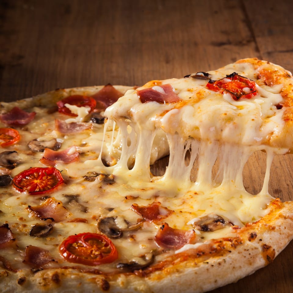 A slice of Pietro's Pizza with melted cheese, bacon, mushrooms, and tomatoes being lifted.